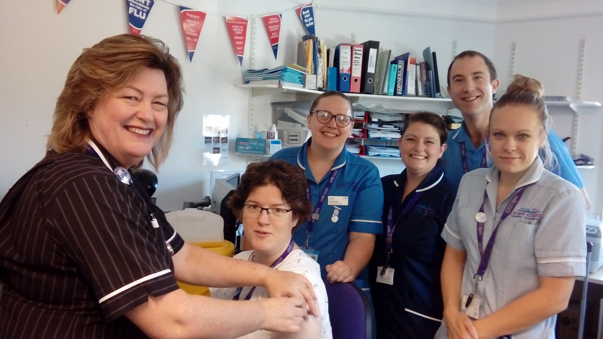 A nurse giving another nurse the flu jab with other staff members qued up